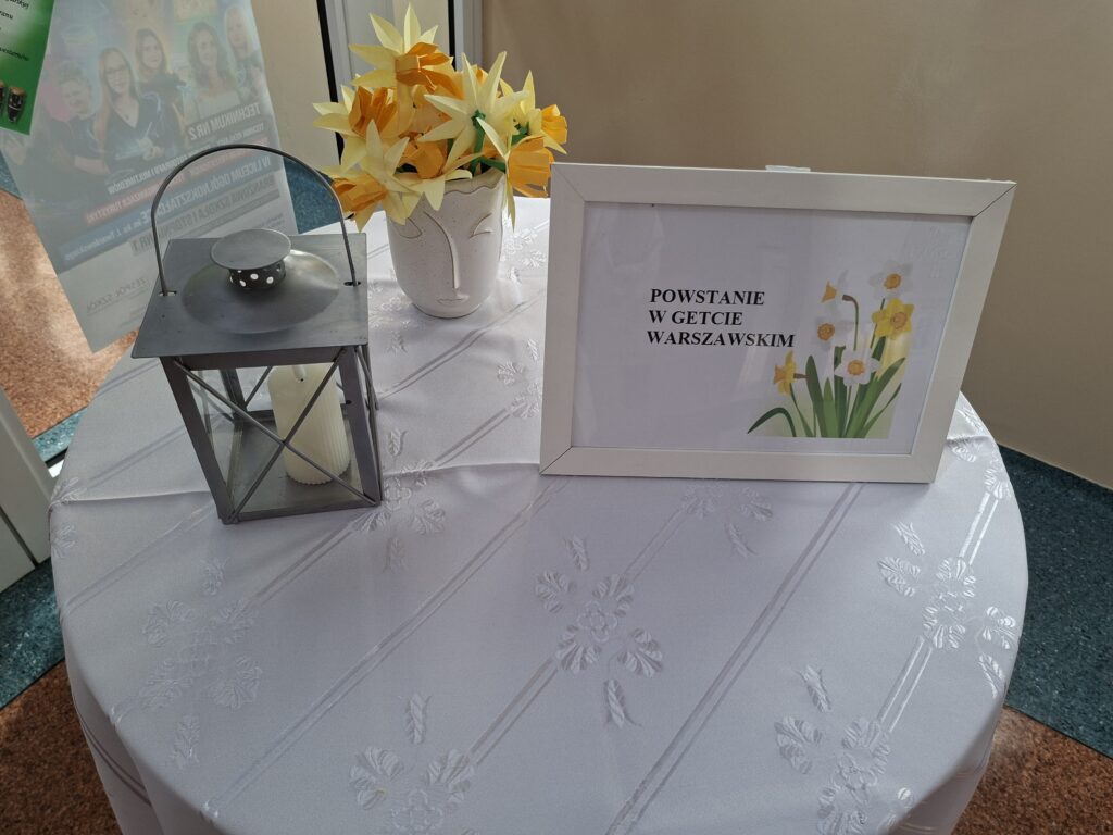Przedstawia stół nakryty białym obrusem, na którym znajduje się szary lampion z białą świeczką oraz ramka w której jest napis "Powstanie w getcie warszawskim" a obok napisu są żółte żonkile.