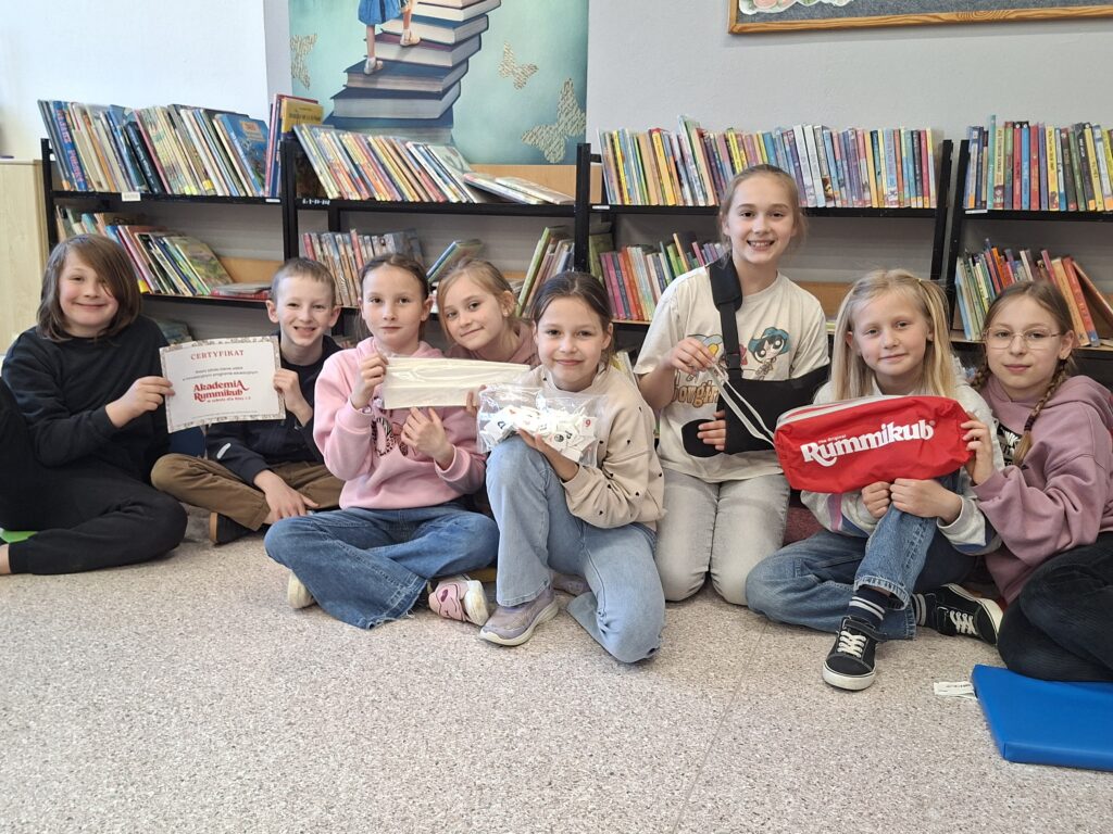Uśmiechnięci uczniowie trzymają w rękach grę "Rummikub" - kolorowe płytki z białymi podstawkami oraz certyfikat uczestnictwa w programie. Uczniowie siedzą na podłodze. W tle regały z książkami.
