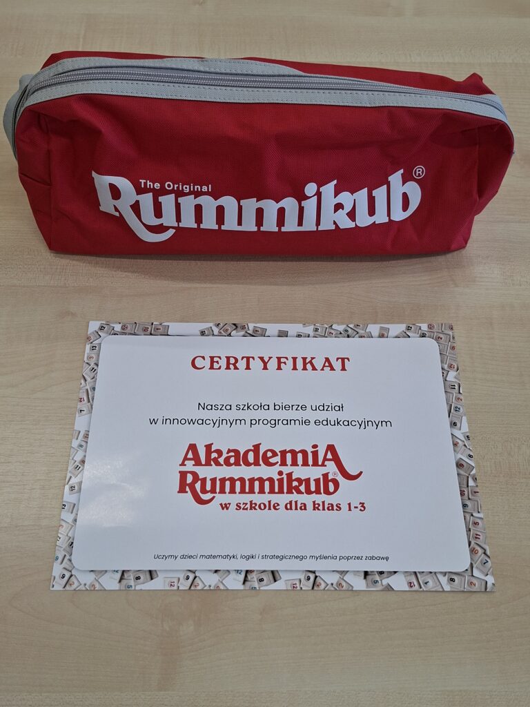 Zdjęcie przedstawia czerwony pokrowiec z białym napisem "The Original Rummikub" oraz certyfikat - ramka składa się z kolorowych płytek z liczbami od 1 do 13, tekst napisany czerwoną i czarną czcionką na białym tle "Nasza szkoła bierze udział w innowacyjnym programie edukacyjnym Akademia Rummikub w szkole dla klas 1-3".