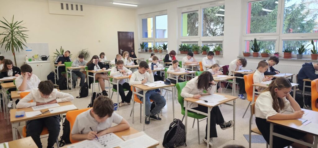  35 edycja Międzynarodowego Konkursu „Kangur Matematyczny”. Na zdjęciu uczniowie ubrani w strój galowy siedzą w klasie przy stolikach i piszą konkurs. 