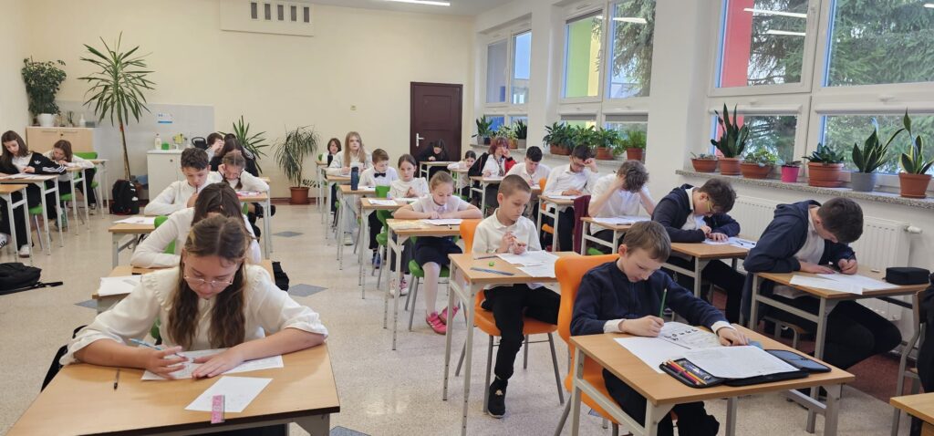  35 edycja Międzynarodowego Konkursu „Kangur Matematyczny”. Na zdjęciu uczniowie ubrani w strój galowy siedzą w klasie przy stolikach i piszą konkurs. 
