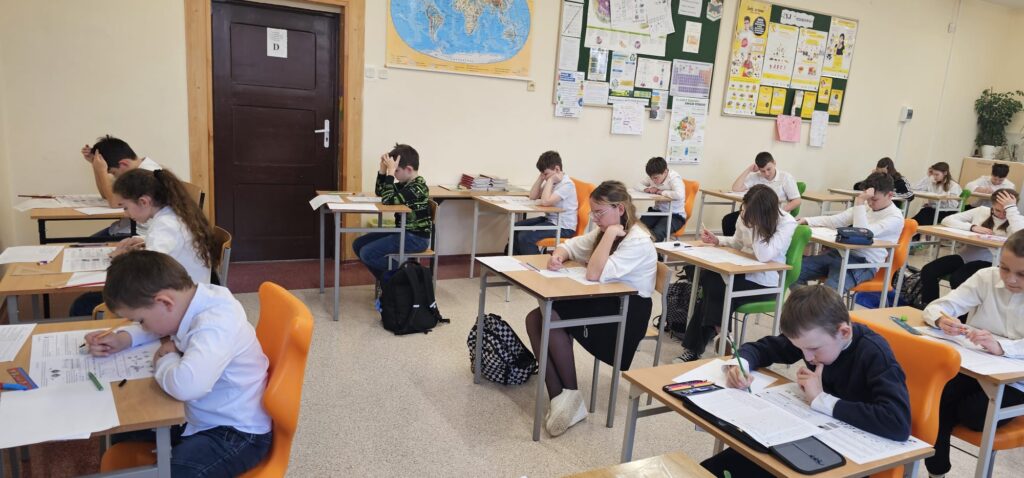  35 edycja Międzynarodowego Konkursu „Kangur Matematyczny”. Na zdjęciu uczniowie ubrani w strój galowy siedzą w klasie przy stolikach i piszą konkurs. 