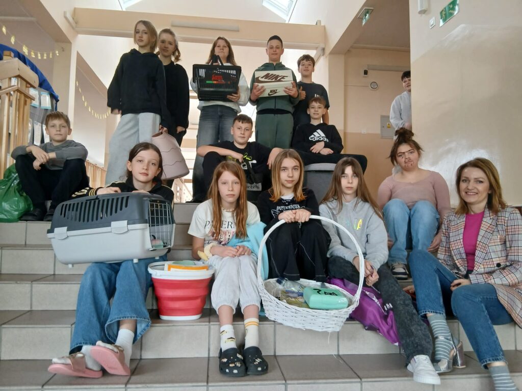 Na zdjęciach uczniowie klas 4–8, zaangażowani w akcję „Dnia bez plecaka” oraz „Dnia Grażynki i Janusza”, siedzą na schodach na korytarzu szkolnym, dumnie prezentując swoje książki w różnych opakowaniach oraz pozując w charakterystycznych sandałach założonych do skarpet.
