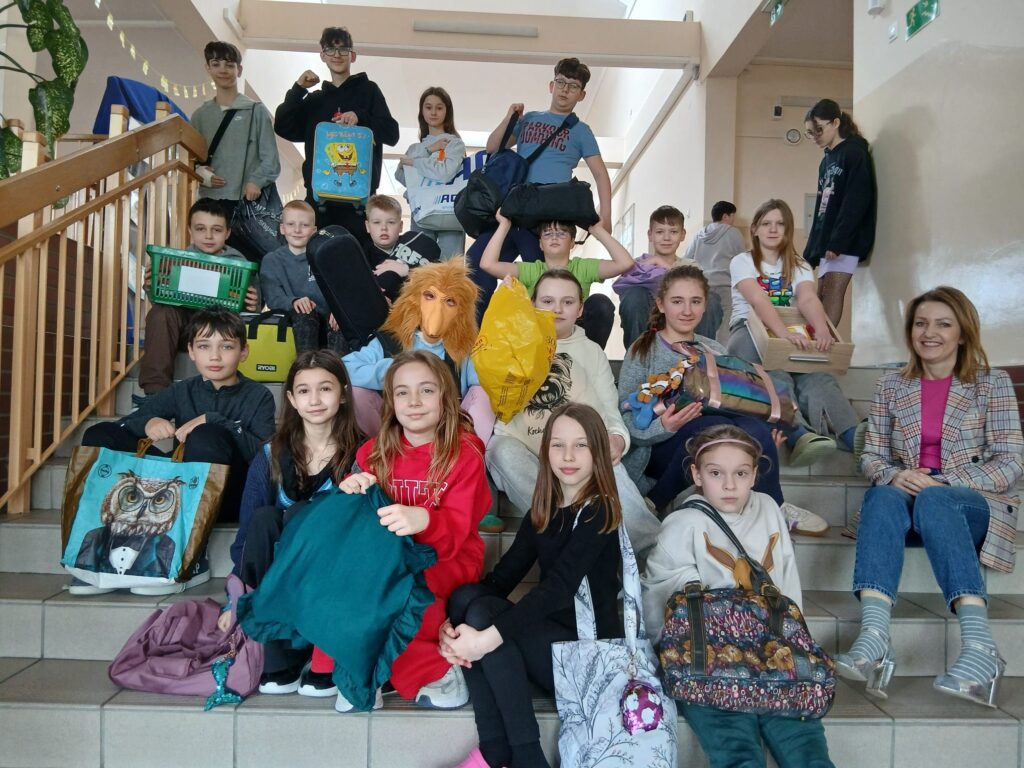 Na zdjęciach uczniowie klas 4–8, zaangażowani w akcję „Dnia bez plecaka” oraz „Dnia Grażynki i Janusza”, siedzą na schodach na korytarzu szkolnym, dumnie prezentując swoje książki w różnych opakowaniach oraz pozując w charakterystycznych sandałach założonych do skarpet.
