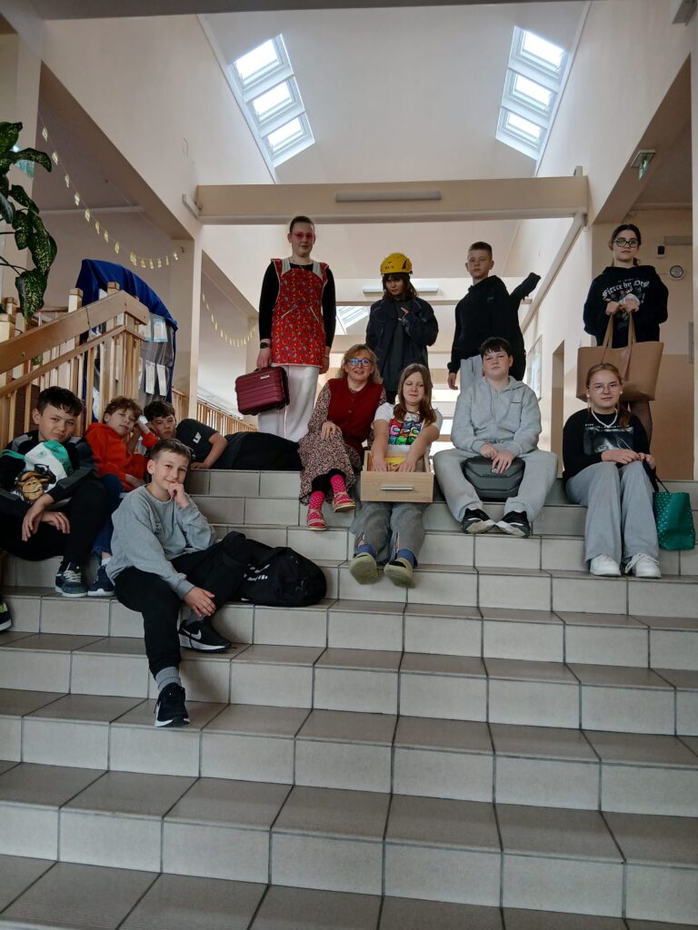 Na zdjęciach uczniowie klas 4–8, zaangażowani w akcję „Dnia bez plecaka” oraz „Dnia Grażynki i Janusza”, siedzą na schodach na korytarzu szkolnym, dumnie prezentując swoje książki w różnych opakowaniach oraz pozując w charakterystycznych sandałach założonych do skarpet.
