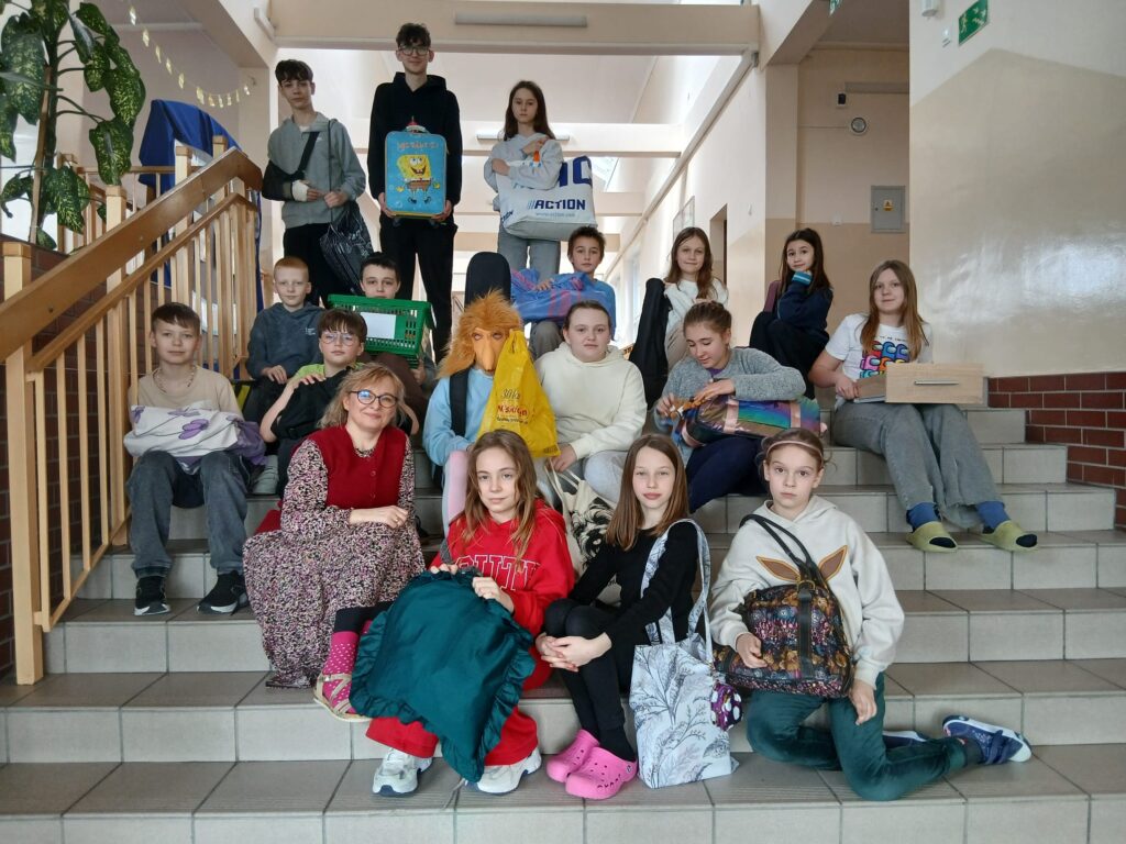 Na zdjęciach uczniowie klas 4–8, zaangażowani w akcję „Dnia bez plecaka” oraz „Dnia Grażynki i Janusza”, siedzą na schodach na korytarzu szkolnym, dumnie prezentując swoje książki w różnych opakowaniach oraz pozując w charakterystycznych sandałach założonych do skarpet.
