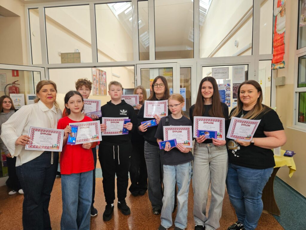 1.Na zdjęciu grupa osób stojąca na korytarzu szkolnym przy szklanym przejściu . Od lewej nauczycielka języka angielskiego  Magdalena Trzcińska w białej koszuli, następnie laureaci konkursu języka angielskiego trzymający dyplomy w rękach dwóch chłopców i pięć dziewczynek. Ostatnia z prawej nauczycielka Sylwia Grzankowska w czarnej koszulce z kolorowym wzorem.