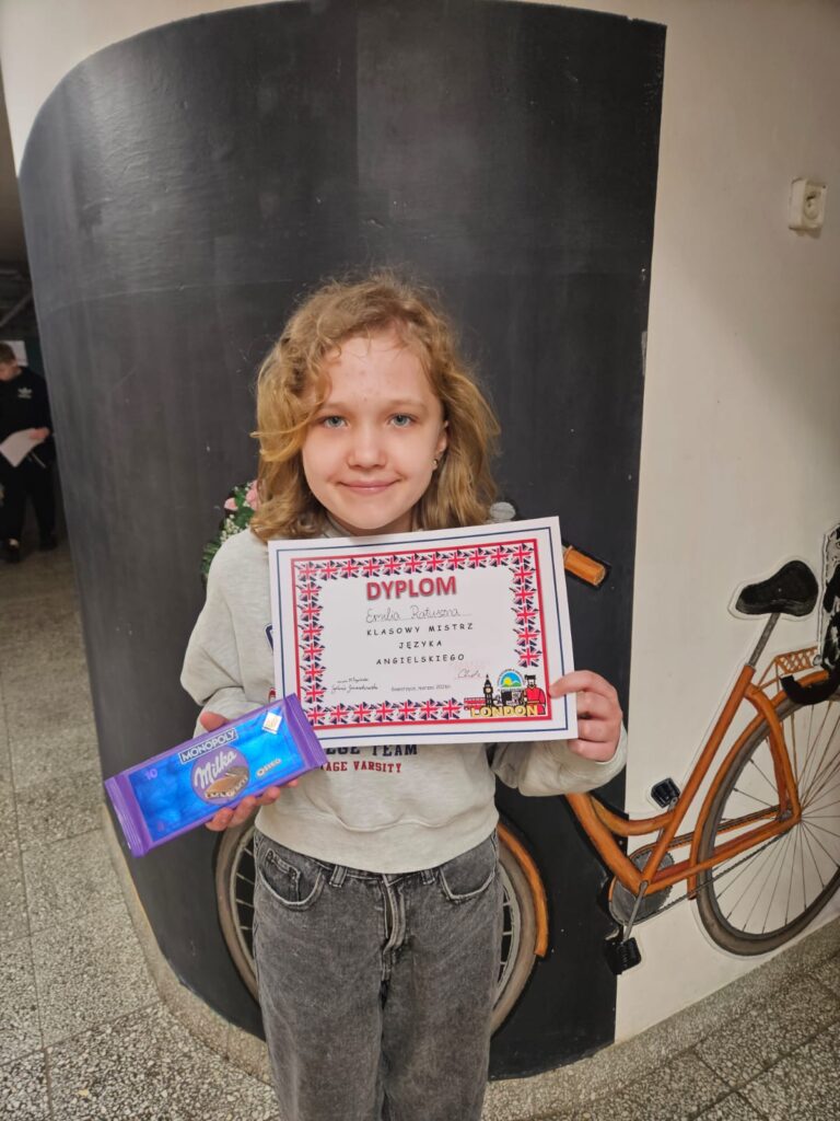 Na zdjęciu uczennica klasy 4b, uśmiechnięta dziewczynka o blond włosach. Stoi na korytarzu szkolnym . W ręku trzyma dyplom z konkursu i czekoladę.
