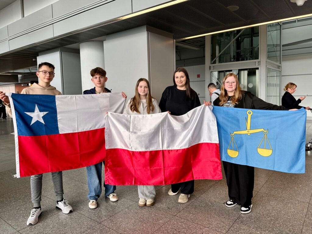 Nasi uczniowie lecą do Chile. Na zdjęciu dwóch uczniów trzyma flagę Chile, opekunkja i jedna z uczennic trzymają flagę Polski, druga uczennica trzyma flagę zgodną z herbem Gminy Gaworzyce (złota waga szalkowa przechylona na lewą stronę na niebeskim materiale). 