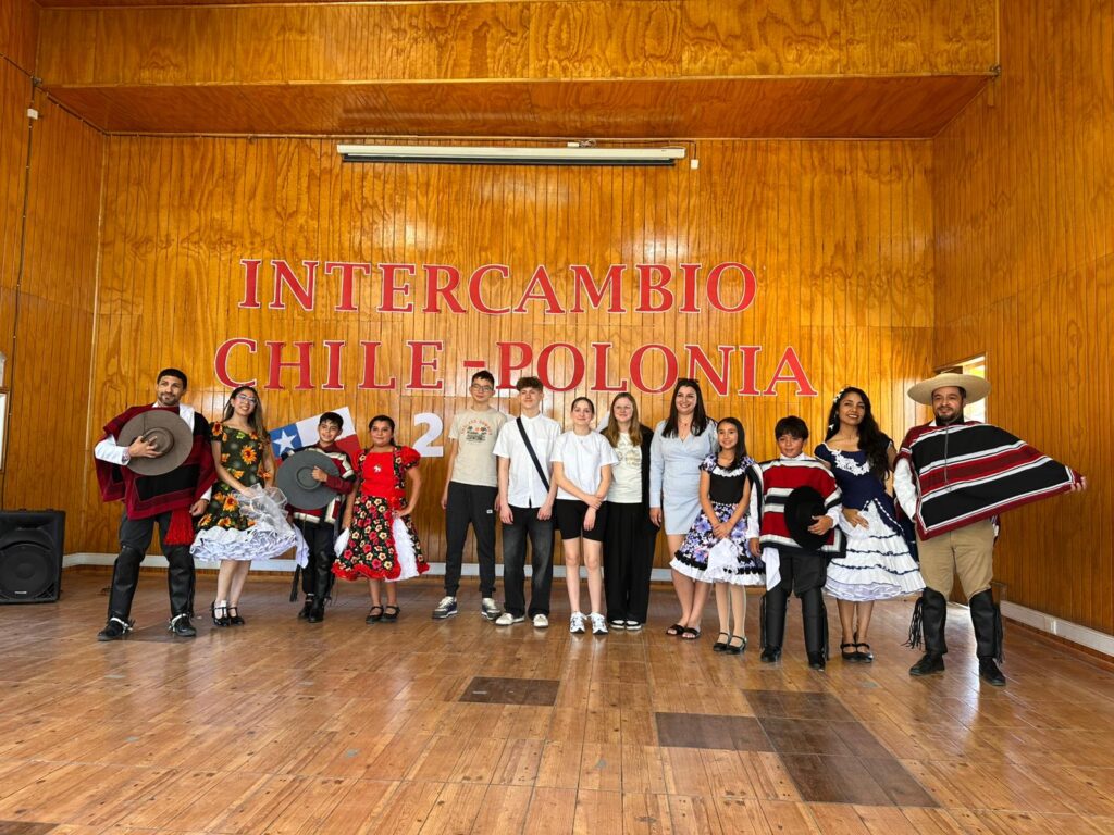 Nasi uczniwoie dotarli do Chile. Na zdjęciu uczniowie z Polski wraz z uczniami z Chile. W tle napis: Intercambio Chile-Polonia. Koło uczniów stoją dorosłe osoby ubrane w narodowe stroje chilijskie. 