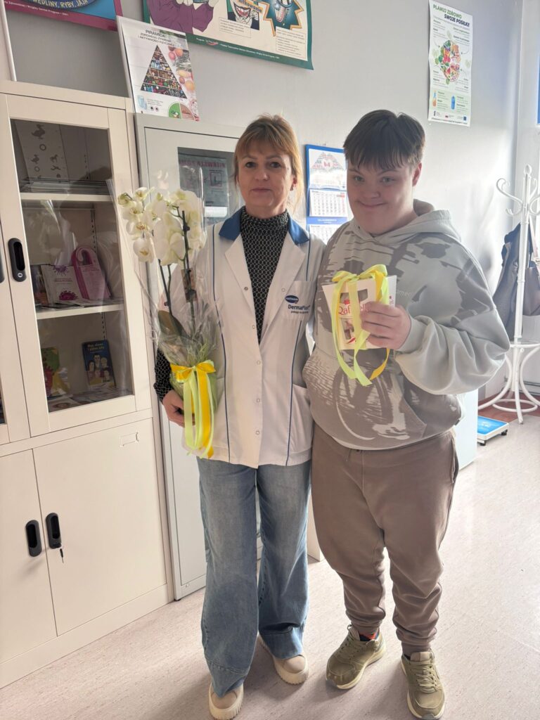 Życzenia z okazji Dnia Służby Zdrowia. Na zdjęciu uczeń wręcza pielęgniarce szkolnej czekoladki. Pielęgniarka w prawej ręce trzyma kremowego storczyka.