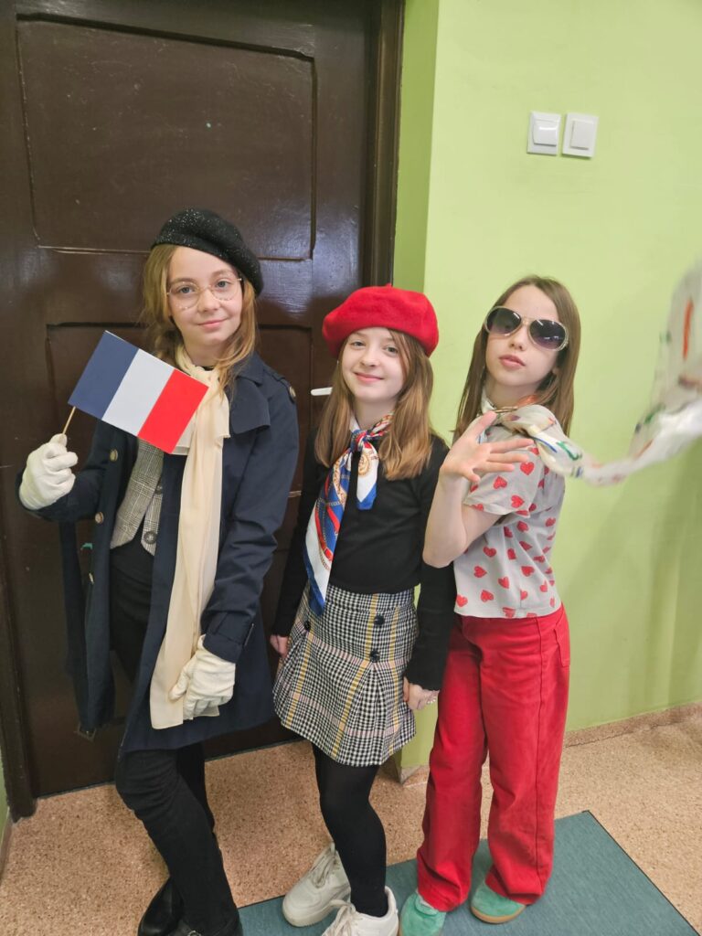 Na zdjęciu trzy dziewczynki .Od lewej: Blondynka w czarnym berecie, ma na sobie czarny płaszcz i beżową apaszkę. W ręce trzyma francuską flagę. Po środku dziewczynka w czerwonym berecie. Ma kolorową apaszkę na szyi, czarną bluzkę i spódniczkę w kratkę. Ostatnia dziewczynka ma okulary przeciwsłoneczne, kolorową bluzkę i apaszkę oraz czerwone spodnie. 