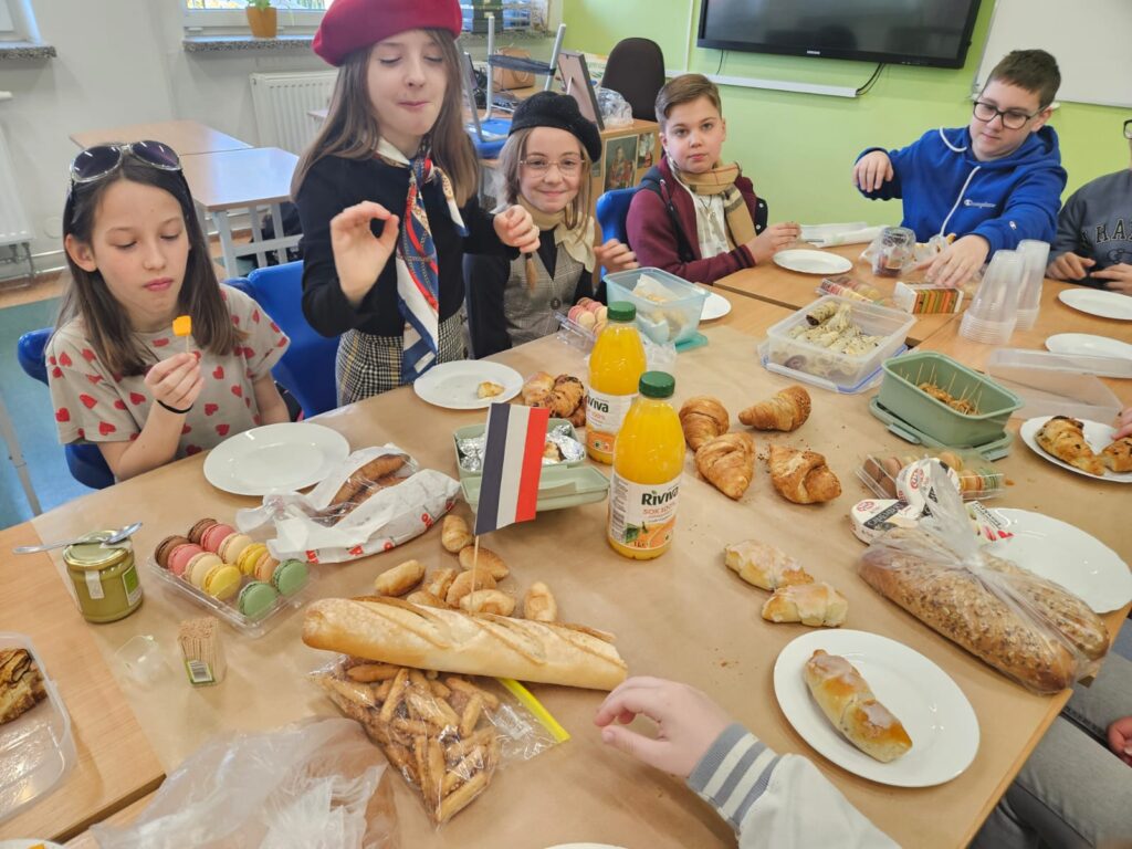 Na pierwszym planie znajduje się duży stół wypełniony różnymi przysmakami kuchni francuskiej. Są tu makaroniki, rogaliki, bagietka, sery i sok pomarańczowy. Dookoła siedzą uczniowie i częstują się jedzeniem.