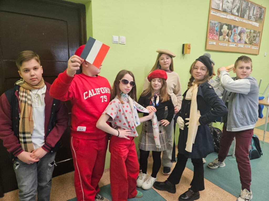 Grupka uczniów stojąca obok drzwi w klasie. Od lewej chłopiec z szalikiem na szyi, drugi jest chłopiec ubrany cały na czerwono, na głowie ma beret a w ręku flagę francuską. Kolejna to dziewczynka z okularach przeciwsłonecznych, w  kolorowej bluzce i apaszce oraz w czerwonych spodniach. Kolejna yo dziewczynka w czerwonym berecie i spódniczce w kratkę. Stoi z ręką na biodrze. Za nią stoi blondynka w okularach i beżowym berecie na głowie. Przedostatnia dziewczynka ma czarny beret i elegancki płaszcz. Na szyi ma beżową apaszkę. Ostatni t chłopiec z bagietką w ręku. Wszyscy zabawnie pozują do zdjęcia.
