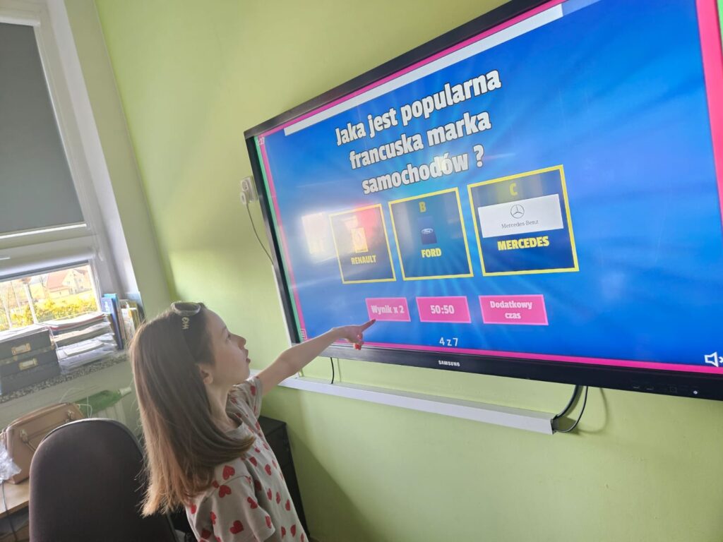 Na zdjęciu dziewczynka stojąca przed tablicą interaktywną. Na tablicy wyświetlone jest pytanie o markę francuskiego samochodu. Dziewczynka zastanawia się nad odpowiedzią.