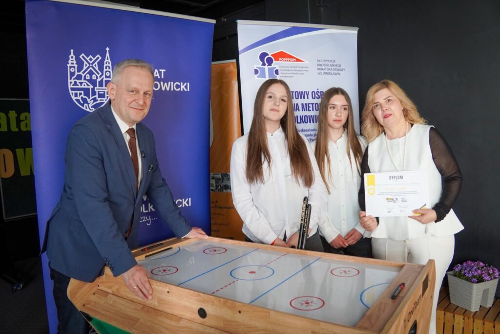 Na zdjęciu widać cztery osoby stojące przy stole do gry przypominającym miniaturowe boisko (np. do air hockeya lub podobnej gry edukacyjnej). Po lewej stronie stoi mężczyzna w eleganckim garniturze – starosta polkowicki. Opiera ręce o stół i lekko się uśmiecha, pozując do zdjęcia. Obok niego stoją dwie młode dziewczyny – uczennice szkoły podstawowej. Są ubrane schludnie, w jasne koszule, i stoją spokojnie obok siebie, patrząc w stronę aparatu. Po prawej stronie znajduje się kobieta – nauczycielka. Trzyma w rękach dyplom, który jest skierowany w stronę aparatu, co sugeruje, że zdjęcie zostało wykonane podczas jakiegoś wydarzenia szkolnego lub konkursu. W tle widać roll-upy (banery) z logotypami instytucji oraz napisami w języku polskim, co wskazuje na oficjalny charakter wydarzenia – prawdopodobnie wręczenie nagrody.