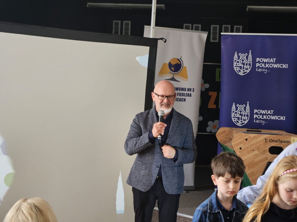 Na zdjęciu widać mężczyznę stojącego przed ekranem projekcyjnym i trzymającego mikrofon, jakby przemawiał do zgromadzonej publiczności. Jest ubrany elegancko – w marynarkę i koszulę – oraz ma okulary i siwą brodę. Wygląda na spokojnego i skupionego.

W tle znajdują się roll-upy (banery) z logo oraz napisami, m.in. „Powiat Polkowicki” oraz grafika przedstawiająca globus i książkę, co sugeruje wydarzenie edukacyjne lub szkolne.

Na pierwszym planie, po prawej stronie, widać dzieci siedzące przy stole lub ławkach. Są skupione na swoich zajęciach i nie patrzą na mężczyznę. Całość sprawia wrażenie spotkania w szkole lub instytucji edukacyjnej, gdzie ktoś prowadzi prezentację lub przemówienie dla młodszej publiczności.
