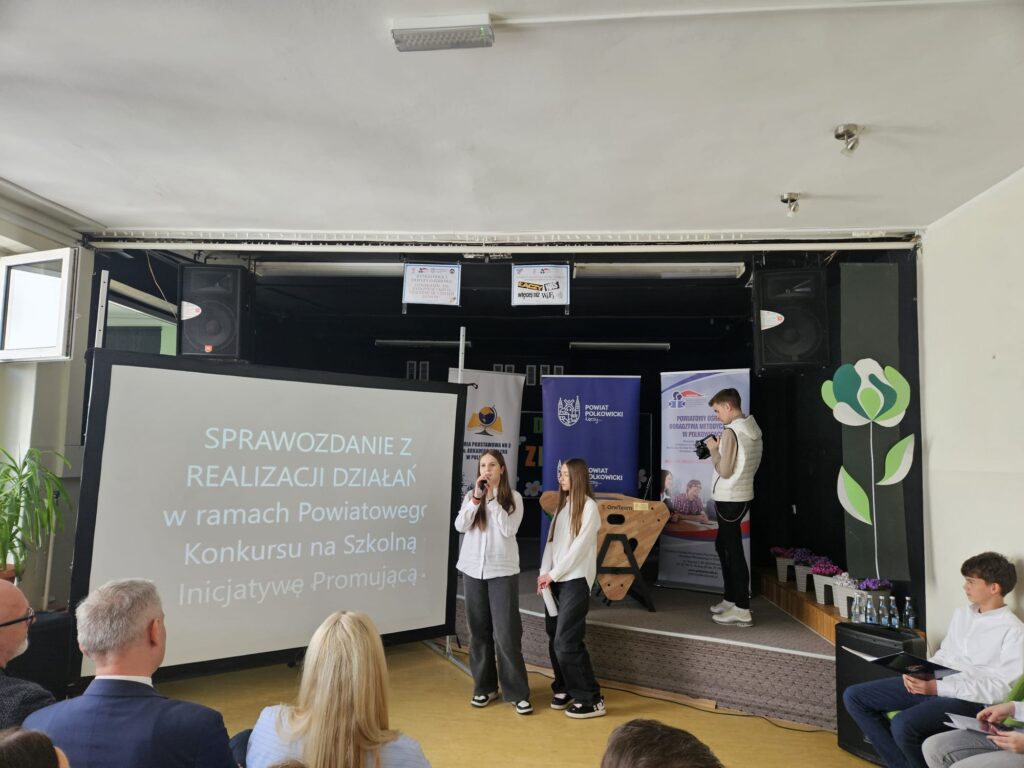 Na zdjęciu widać scenę w sali przypominającej szkolną aulę lub świetlicę, gdzie odbywa się prezentacja lub wystąpienie. Na środku stoją dwie dziewczyny – jedna z nich trzyma mikrofon i mówi, druga stoi obok, słuchając lub przygotowując się do swojej części. Obie są ubrane raczej schludnie, w jasne koszule i ciemne spodnie.

Po lewej stronie znajduje się duży ekran projekcyjny z wyświetlonym tytułem prezentacji: „Sprawozdanie z realizacji działań w ramach Powiatowego Konkursu na Szkolną Inicjatywę Promującą…”, co wskazuje na oficjalne wydarzenie szkolne lub konkursowe.

W tle stoją banery, m.in. z napisem „Powiat Polkowicki”, co sugeruje, że wydarzenie jest organizowane lub wspierane przez lokalne władze. Po prawej stronie na podwyższeniu stoi chłopak, który wygląda, jakby przygotowywał sprzęt lub robił zdjęcia. Obok niego widać dekoracje roślinne i kilka butelek ustawionych na stoliku.

Na pierwszym planie siedzą dorośli widzowie – prawdopodobnie nauczyciele, rodzice lub goście – obserwujący wystąpienie. Całość przedstawia formalne, szkolne wydarzenie z udziałem uczniów i publiczności.