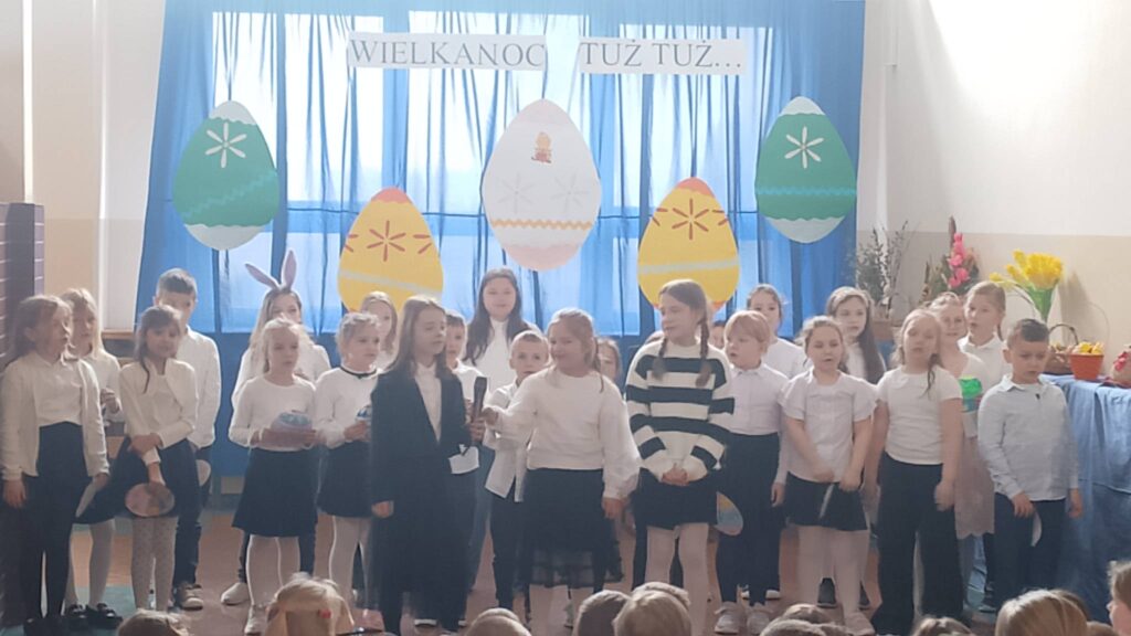 Apel wielkanocny. Uczniowie ubrani w strój galowy stoją na tle dekoracji świątecznej: pięć dużych kolorowych pisanek wisi na tle niebieskiego materiału, nad pisankami napis Wielkanoc tuż tuż. Uczniwoie biorą udział w apelu, recytują wierszyki i śpiewają piosenki.