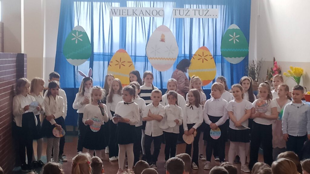 Apel wielkanocny. Uczniowie ubrani w strój galowy stoją na tle dekoracji świątecznej: pięć dużych kolorowych pisanek wisi na tle niebieskiego materiału, nad pisankami napis Wielkanoc tuż tuż. Uczniwoie biorą udział w apelu, recytują wierszyki i śpiewają piosenki.