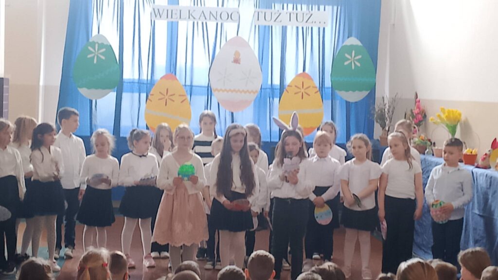 Apel wielkanocny. Uczniowie ubrani w strój galowy stoją na tle dekoracji świątecznej: pięć dużych kolorowych pisanek wisi na tle niebieskiego materiału, nad pisankami napis Wielkanoc tuż tuż. Uczniwoie biorą udział w apelu, recytują wierszyki i śpiewają piosenki.