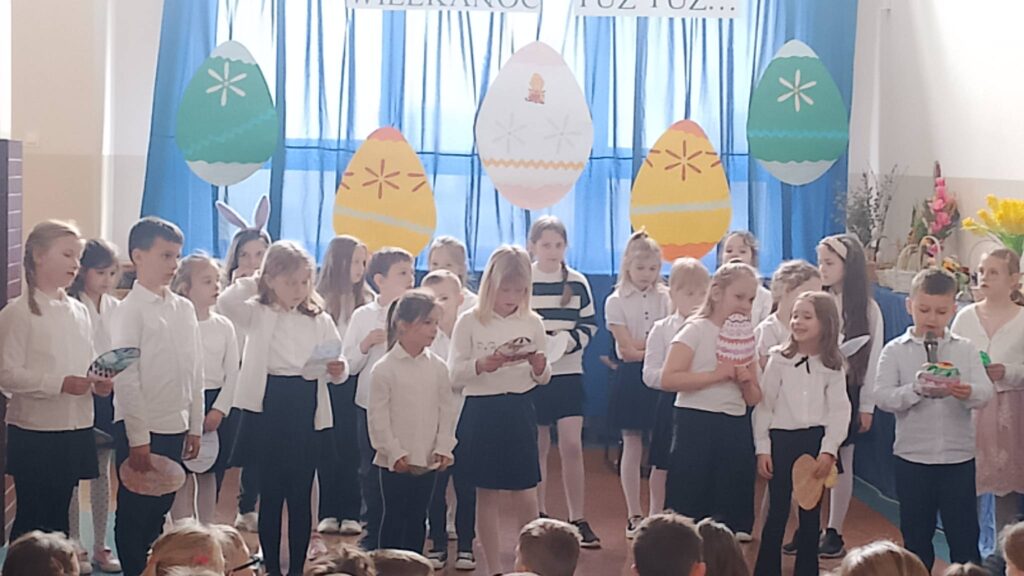 Apel wielkanocny. Uczniowie ubrani w strój galowy stoją na tle dekoracji świątecznej: pięć dużych kolorowych pisanek wisi na tle niebieskiego materiału, nad pisankami napis Wielkanoc tuż tuż. Uczniwoie biorą udział w apelu, recytują wierszyki i śpiewają piosenki.