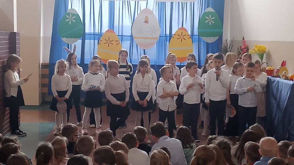 Apel wielkanocny. Uczniowie ubrani w strój galowy stoją na tle dekoracji świątecznej: pięć dużych kolorowych pisanek wisi na tle niebieskiego materiału, nad pisankami napis Wielkanoc tuż tuż. Uczniwoie biorą udział w apelu, recytują wierszyki i śpiewają piosenki.