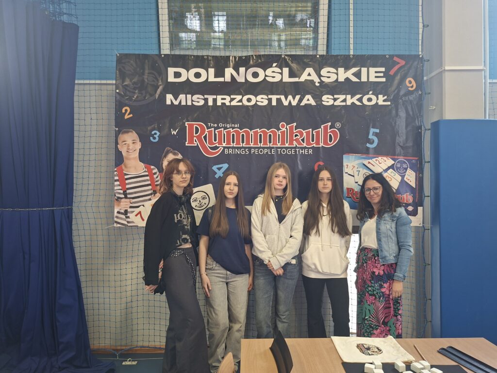 Zdjęcie przedstawia uczennice szkoły stojące obok siebie wraz z nauczycielem. Nad uczniami i nauczycielem wisi biało-niebieski napis Dolnośląskie Mistrzostwa Szkół na niebieskim tle. Osoby te stoją obok niebieskiej kotary, która przedziela salę gimnastyczną.