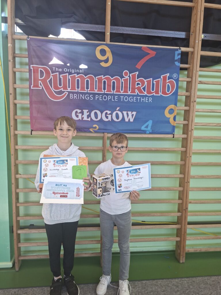 Zdjęcie przedstawia dwóch chłopców - laureatów konkursu trzymających w rękach dyplomy oraz nagrody - "Rummikub słowny". W tle drewniane drabinki. Nad chłopcami wisi granatowa flaga z kolorowym napisem Rummikub.