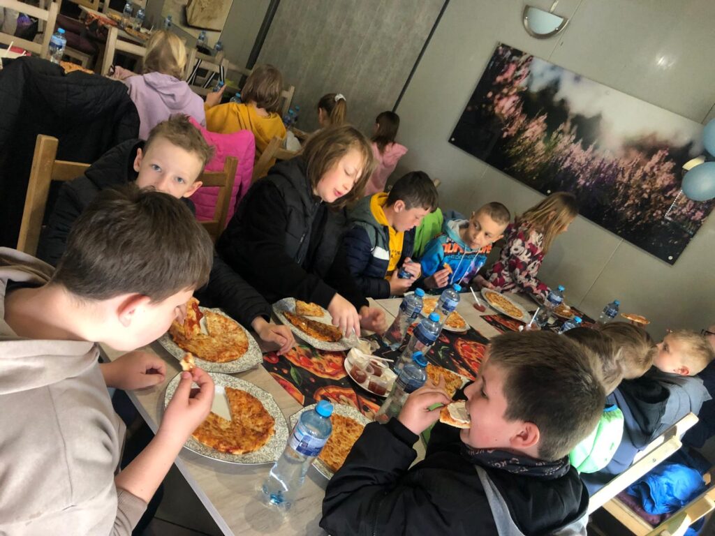 Grupa uczniów podczas posiłku po wycieczce krajoznawczej, uczniwoie jedzą pizzę, w tle obraz wrzosami. 