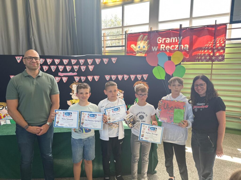 Zdjęcie przedstawia uczniów szkoły (zwycięską drużynę) trzymających dyplomy i nagrodę - grę "Ubongo" oraz zielony medal za zajęcie 1 miejsca. W tle kolorowa dekoracja z balonami.