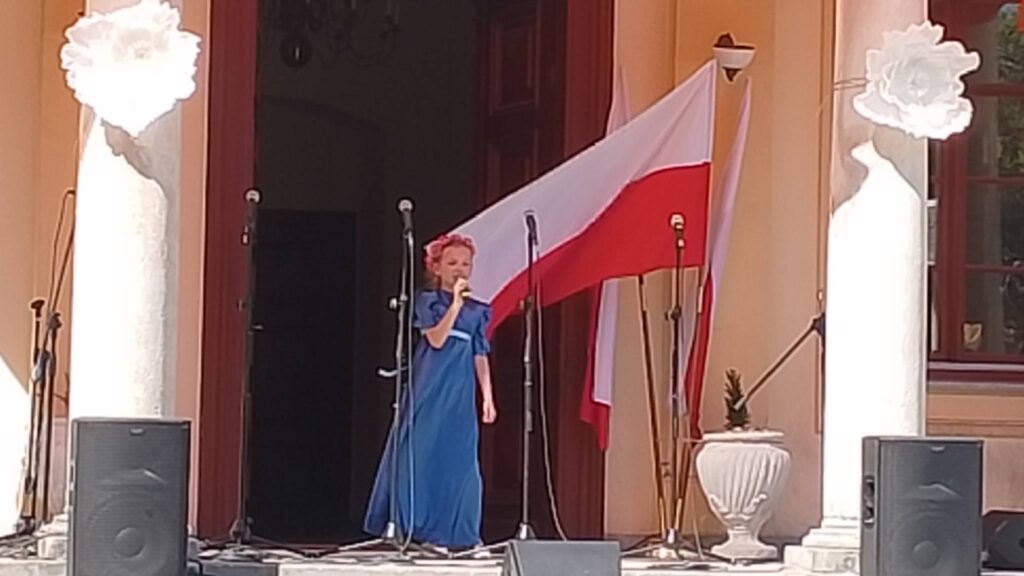 Obchody rocznicy uchwalenia Konstytucji 3 Maja. Na zdjęciu uczennica klasy trzeciej ubrana w długą chabrową sukienkę stoi na tle drewnianych drzwi od pałacu i śpiewa piosenkę. 