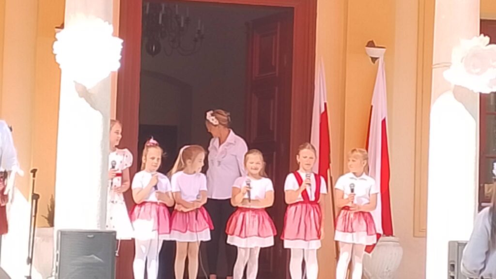 Obchody rocznicy uchwalenia Konstytucji 3 Maja. Uczennice z klas 1-3 ubrane w białe bluzeczki i biało-czerwone spódniczki stoją na tle drzwi od pałacu, trzymają mikrofon i czekają na wykonanie piosenki. 