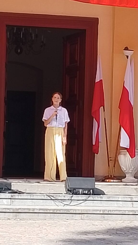 Obchody rocznicy uchwalenia Konstytucji 3 Maja. Na zdjeciun uczennica klasy piątej ubrana w  białą elegancką  bluzkę i żółte spodnie stoi na tle drewnianych drzwi od pałacu, trzyma mikrofon i wykonuje piosenkę. 