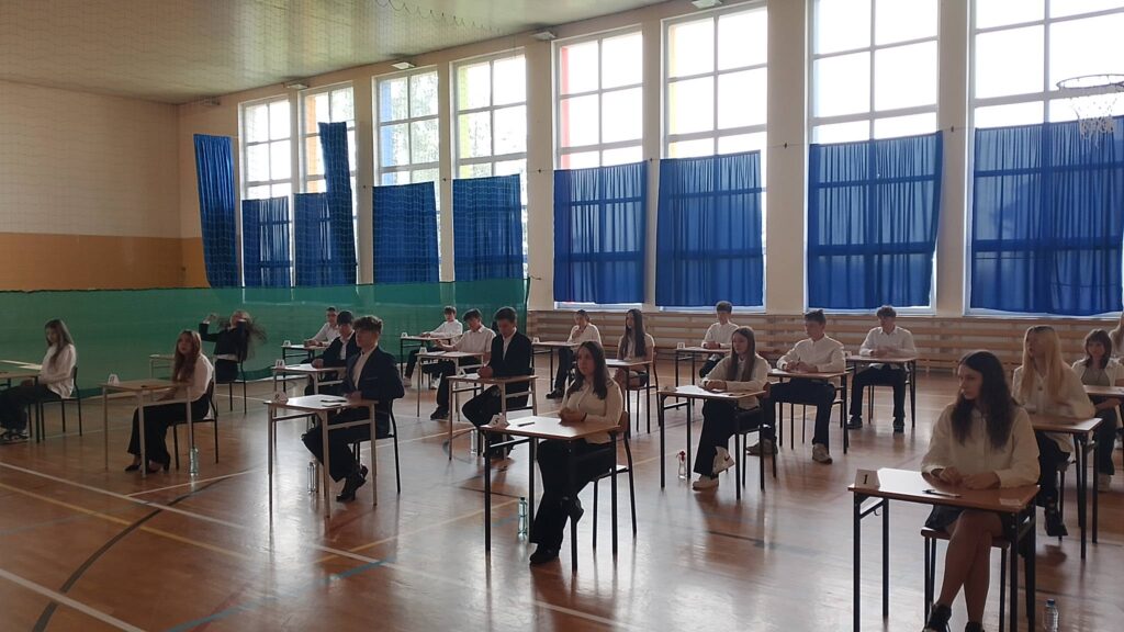 Egzamin ósmoklasisty. Na sali 20 uczniów klasy ósmej ubranych na galowo siedzi przy ławeczkach na sali gimnastycznej czekając na rozdanie arkuszy egzminacyjnych. 