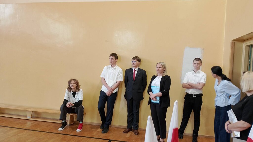 Egzamin ósmoklasisty. Na sali 4 uczniów klasy ósmej ubranych na galowo stoi na sali gimnastycznej czekając na rozdanie arkuszy egzminacyjnych. 
