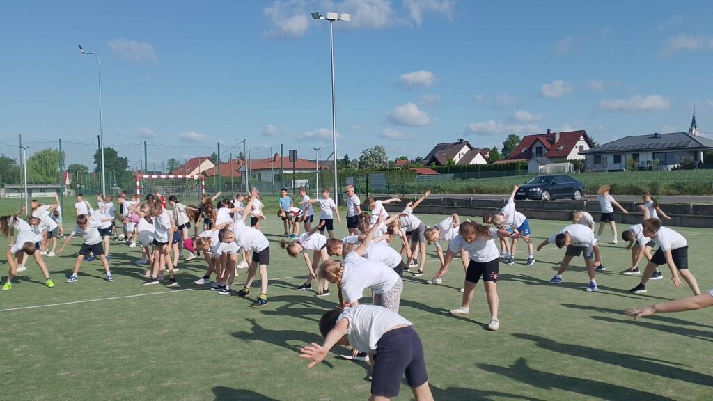 Na zdjęciu widać dużą grupę uczniów uczestniczących w porannej rozgrzewce na szkolnym boisku. Dzieci są ubrane w stroje sportowe – głównie białe koszulki i ciemne spodenki – i wykonują jednocześnie ćwiczenia rozciągające. Większość z nich pochyla się na boki z wyciągniętymi ramionami, ćwicząc gibkość i przygotowując ciało do dalszej aktywności fizycznej.

Zajęcia odbywają się na zielonej nawierzchni boiska sportowego w słoneczny dzień. Długie cienie rzucane przez uczestników świadczą o porannych godzinach lub późnym popołudniu. W tle widoczna jest bramka do piłki ręcznej, wysokie ogrodzenie boiska oraz latarnie oświetleniowe.