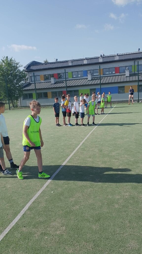 Na zdjęciu przedstawiono grupę dzieci uczestniczących w zajęciach sportowych na szkolnym boisku. Uczniowie stoją w kilku rzędach wzdłuż wyznaczonej linii, przygotowując się do kolejnej konkurencji lub ćwiczenia. Część z nich ma na sobie jaskrawozielone znaczniki sportowe, które pomagają rozróżnić drużyny.

Na pierwszym planie znajduje się chłopiec w zielonej kamizelce i sportowych butach, który stoi przy linii startu i wygląda na gotowego do rozpoczęcia zadania. Pozostałe dzieci ustawione są za nim, oczekując na swoją kolej. W tle można dostrzec osobę dorosłą nadzorującą przebieg zajęć.