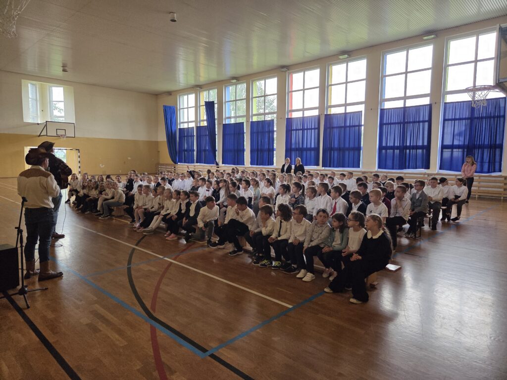 Zdjęcie z koncertu “Kowboje, gitara i banjo”. Przedstawia uczniów klas 1-3 zgromadzonych w sali gimnastycznej, gotowych do uczestniczenia w koncercie. Uczniowie są ubrani w stroje apelowe, siedzą na drewnianych ławkach.
