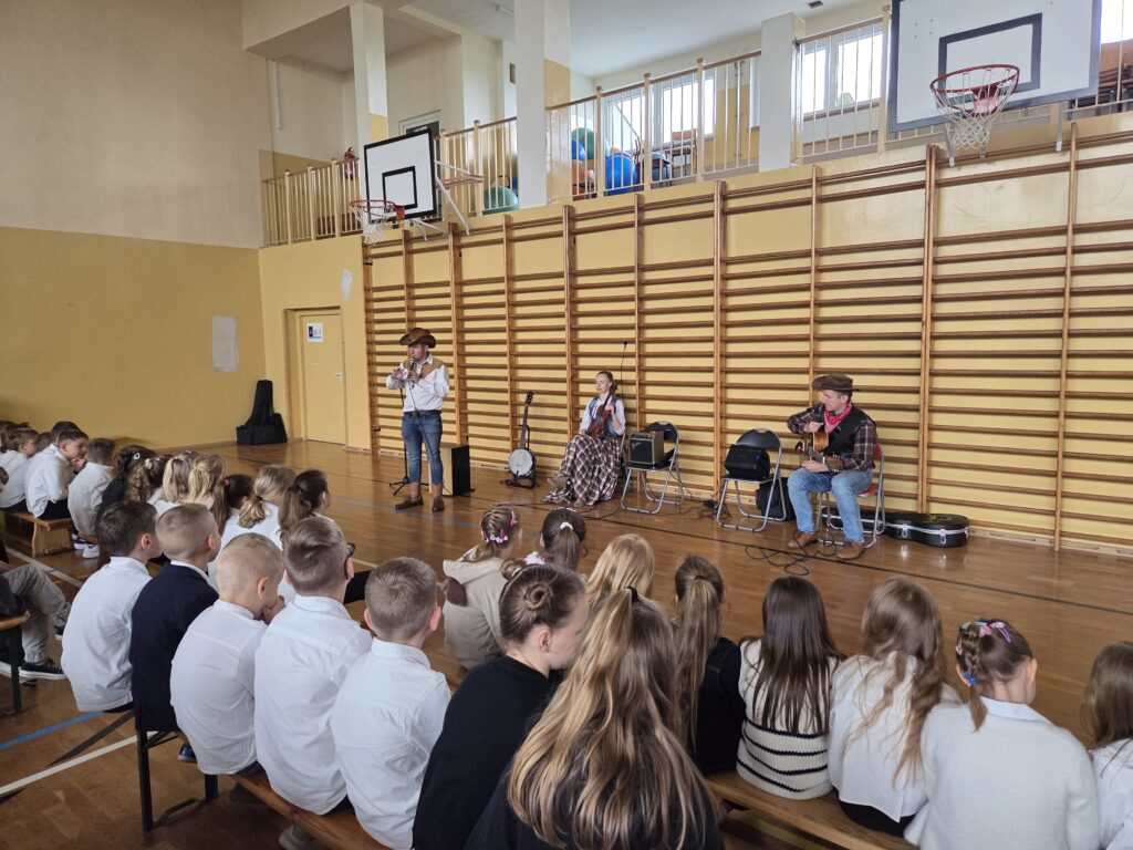 Zdjęcie z koncertu “Kowboje, gitara i banjo”. Przedstawia uczniów klas 1-3 zgromadzonych w sali gimnastycznej, gotowych do uczestniczenia w koncercie. Uczniowie są ubrani w stroje apelowe, siedzą na drewnianych ławkach.
