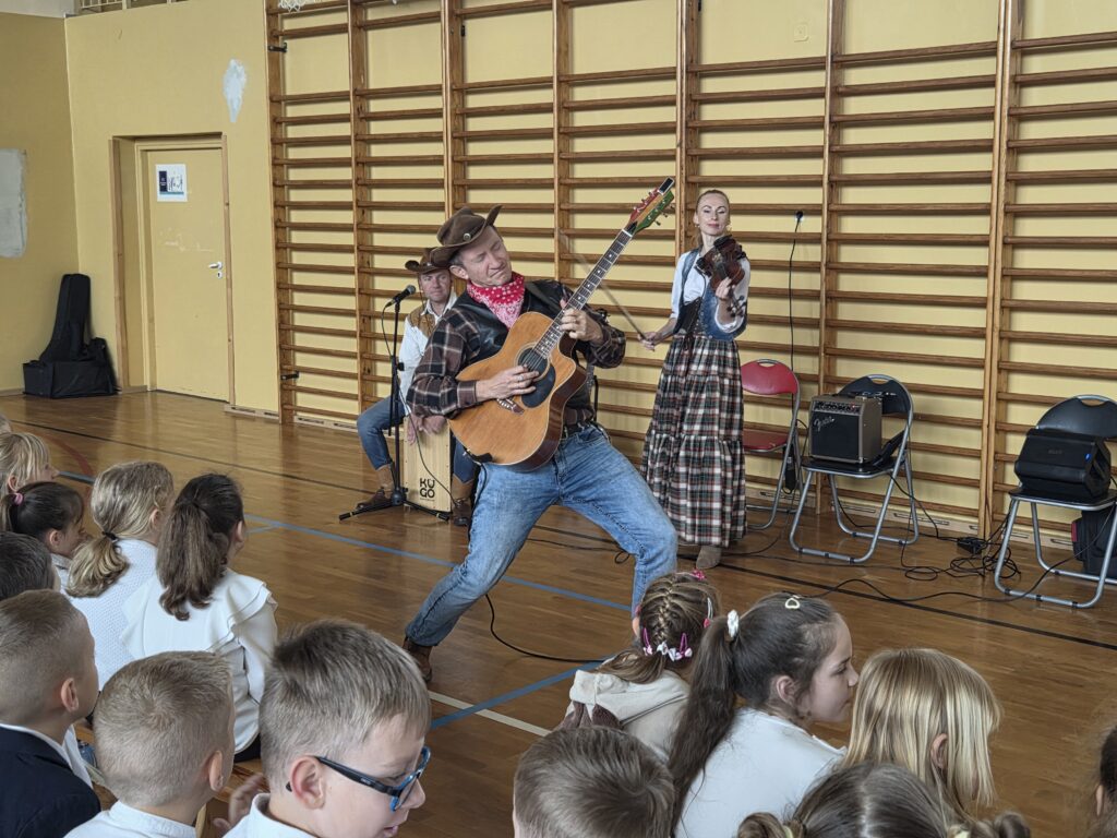 Zdjęcie z koncertu “Kowboje, gitara i banjo”. To zbliżenia na muzyka w stroju kowboja, grającego z pasją na gitarze. W tle kobieta w spódnicy w kratę gra na skrzypcach.
