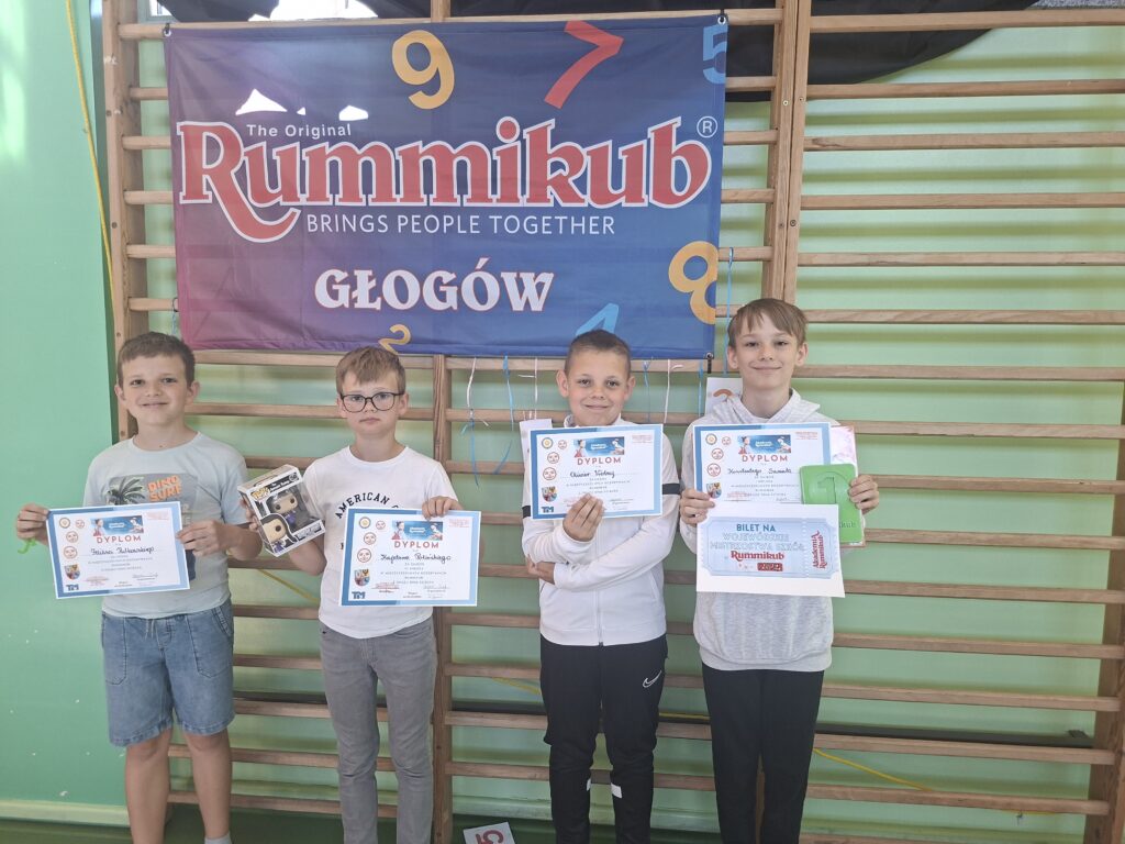 Zdjęcie przedstawia laureatów turnieju z dyplomami i nagrodami oraz "złotym biletem" na Mistrzostwa Wojewódzkie Szkół. Uczniowie są uśmiechnięci. W tle drewniane drabinki i niebieska flaga z kolorowym napisem Rummikub.