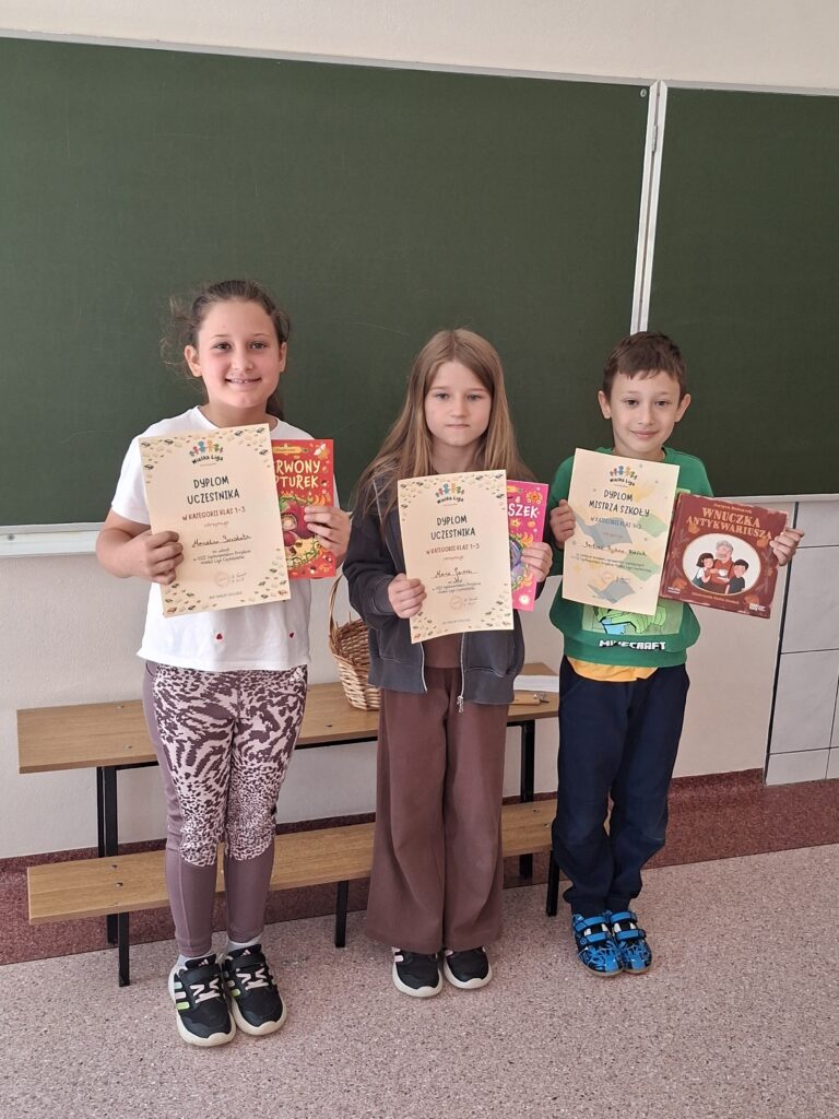 Zdjęcie przedstawia 3 nagrodzonych uczniów trzymających książki i dyplomy w konkursie Wielka Liga Czytelników.  Uczniowie są uśmiechnięci i stoją w klasie. W tle zdjęcia znajduje się zielona tablica.