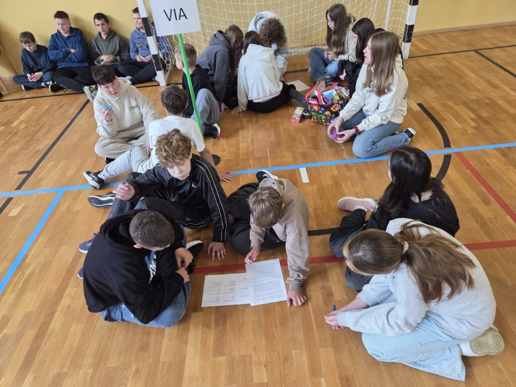 Przedstawia uczniów rozwiązujących łamigłówki w sali gimnastycznej. Uczniowie siedzą lub leżą na parkiecie, trzymają w ręce długopisy i zaznaczają prawidłowe odpowiedzi na kartach pracy. W tle widać bramkę do piłki nożnej.