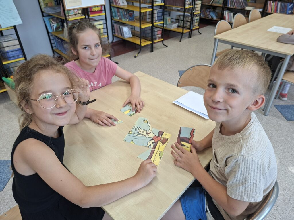 Troje uczniów siedzi przy ławce w bibliotece szkolnej i układają puzzle o Koziołku Matołku. W tle regały z książkami.