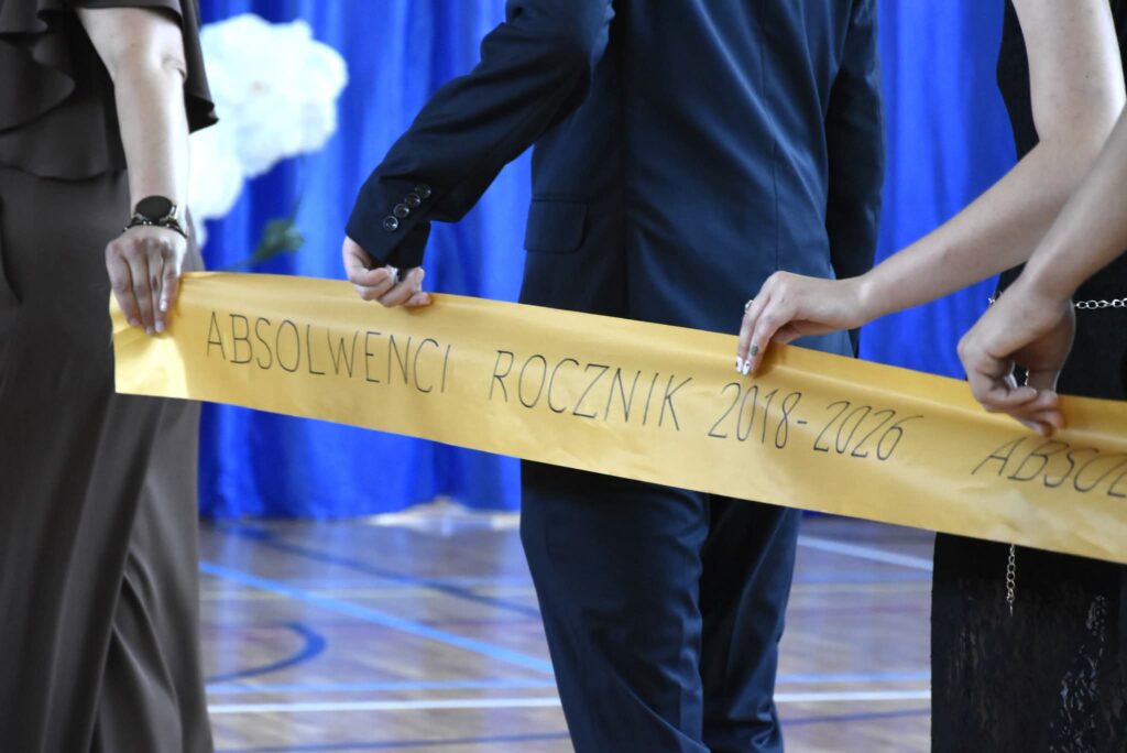 Na zdjęciu widać fragment uroczystości zakończenia szkoły lub balu absolwentów. W centrum kadru znajdują się trzy osoby ubrane w eleganckie stroje – jedna w granatowym garniturze, a dwie w długich, wizytowych sukienkach. Widać je od ramion w dół, więc ich twarze nie są pokazane.

Osoby wspólnie trzymają szeroką, złotą wstęgę z ręcznie napisanym napisem: „ABSOLWENCI ROCZNIK 2018–2026”.