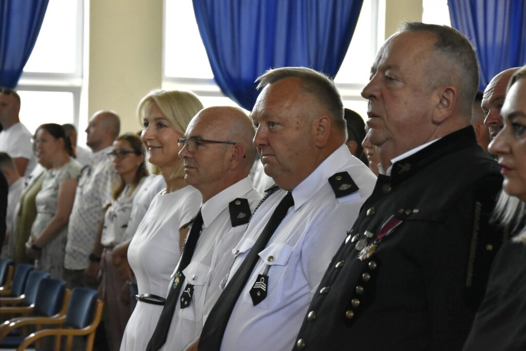 Na zdjęciu przedstawiono uczestników uroczystości szkolnej stojących podczas oficjalnej części wydarzenia. W pierwszym planie znajduje się kilka osób ustawionych w jednym rzędzie. Dwie z nich mają na sobie białe mundury galowe z czarnymi krawatami i oznakami służbowymi, a obok stoi mężczyzna w czarnym mundurze z odznaczeniami przypiętymi do piersi. Wśród nich widoczna jest również kobieta ubrana w elegancką białą sukienkę. W tle znajdują się pozostali uczestnicy uroczystości, stojący między rzędami krzeseł