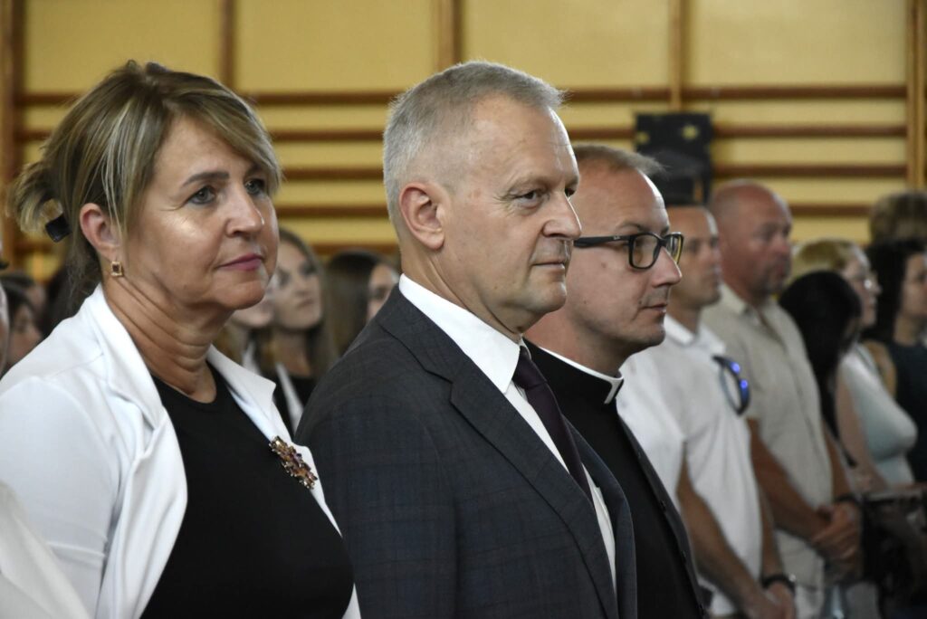 Na zdjęciu przedstawiono uczestników uroczystej akademii szkolnej stojących podczas oficjalnej części wydarzenia. W pierwszym planie widoczne są trzy osoby. Po lewej stronie znajduje się kobieta ubrana w czarną sukienkę i białą marynarkę, ozdobioną elegancką broszką. Obok niej stoi mężczyzna w ciemnoszarym garniturze, białej koszuli i ciemnym krawacie. Po prawej stronie widoczny jest duchowny w czarnym stroju z koloratką oraz okularami.

W tle znajdują się pozostali uczestnicy uroczystości – uczniowie i zaproszeni goście – ustawieni w rzędach. Wszyscy zwróceni są w tym samym kierunku, co sugeruje, że uczestniczą w podniosłym momencie, takim jak odśpiewanie hymnu państwowego, przemówienie lub inny oficjalny punkt programu.