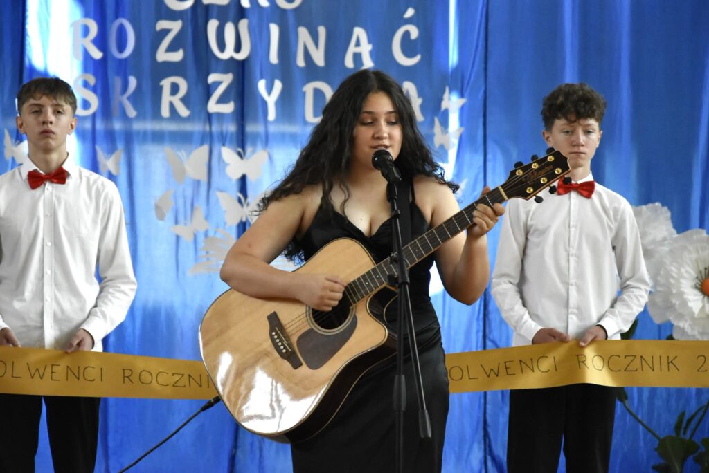 Na zdjęciu uwieczniono występ artystyczny podczas uroczystości szkolnej. W centrum kadru znajduje się uczennica ubrana w elegancką czarną suknię, która śpiewa do mikrofonu i jednocześnie gra na gitarze akustycznej. Jej występ stanowi główny punkt programu.

Po obu stronach wokalistki stoją dwaj uczniowie ubrani w białe koszule, czarne spodnie oraz czerwone muszki. Trzymają oni złotą wstęgę z napisem „Absolwenci Rocznik 2018–2026”, nawiązującą do ceremonii pożegnania absolwentów.