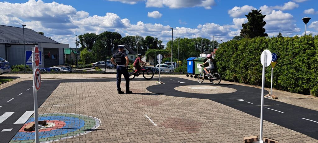 Na zdjęciu przedstawiono praktyczne zajęcia z bezpieczeństwa ruchu drogowego odbywające się na szkolnym miasteczku ruchu drogowego. W centrum kadru stoi policjant, który obserwuje i nadzoruje uczniów poruszających się na rowerach.
Dwoje dzieci w kaskach rowerowych jedzie po wyznaczonej trasie, ćwicząc zasady bezpiecznego poruszania się po drogach. Wokół znajdują się elementy infrastruktury drogowej, takie jak przejście dla pieszych, znaki drogowe, rondo oraz oznakowane jezdnie, które odwzorowują rzeczywiste warunki ruchu.